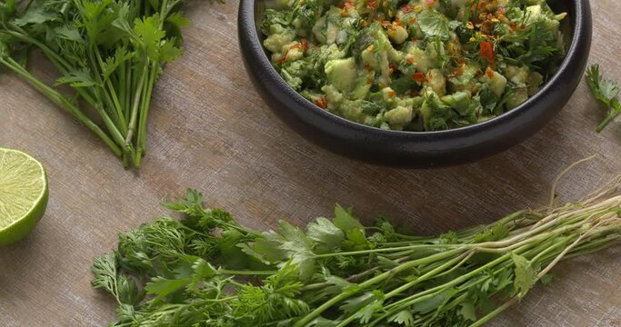 A bowl of homemade guacamole with a lot of chopped fresh coriander, topped with hot chili flakes. Freshly harvested coriander with and without roots on the side. Table spin.