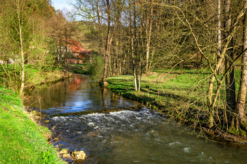 Romantischer Flusslauf mit kleiner Staustufe und rotem Haus in der Fränkischen Schweiz, Deutschland