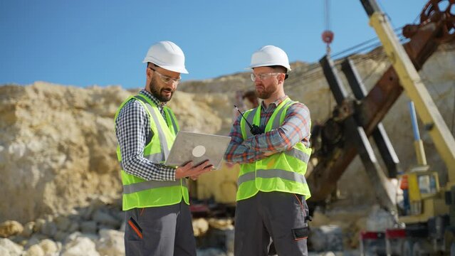 Two engineers geologists looks a quarry excavation plan in laptop, excavator in the background