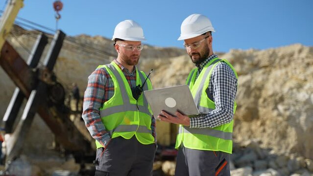Two engineers geologists looks a quarry excavation plan in laptop, excavator in the background
