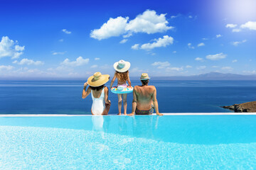 A holiday family sits at the edge of an infinity pool and enjoys the summer day view of the mediterranean sea