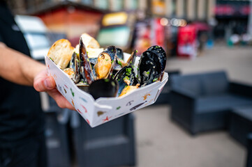 Hand holding a paper container of cooked mussels against a blurred street background, suggesting...