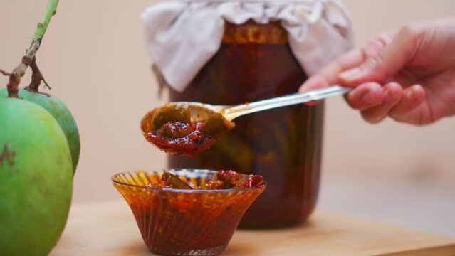 Close up shot of Homemade Mango Pickle or aam ka achar in bowl with raw mangoes and transparent jar in background. Indian homemade raw mango pickle. Indian woman hand with spoon of pickle. 