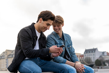 Denmark, Copenhagen, two young men sitting on a bench using cell phone