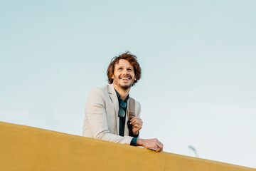 Portrait of laughing businessman with backpack leaning on wall looking at distance