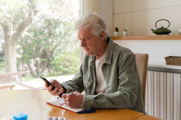Senior diabetic man using mobile phone while writing in diary at home