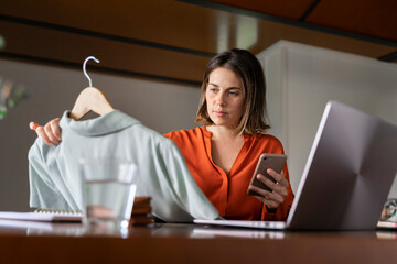 Businesswoman with smart phone looking at clothes while sitting at home