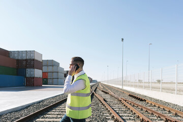Man on railway tracks in front of cargo containers talking on cell phone