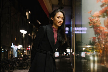 Smiling young woman looking in shop window at night, Frankfurt, Germany