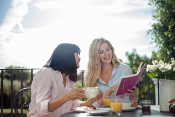Two women having breakfast at outdoor cafe table, reading book