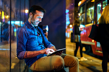 Man with headphones sitting at a station at night using his digital tablet