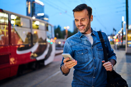 Man using smartphone and listening to music while standing at tram stop in the evening