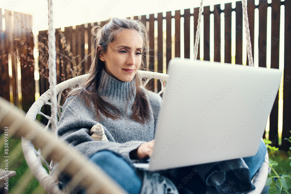 Poster Beautiful young woman using laptop while sitting on swing in garden