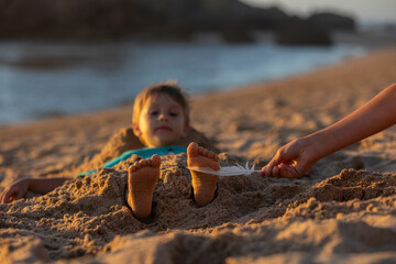 Child, tickling sibling on the beach on the feet with feather, kid cover in sand, smiling, laughing