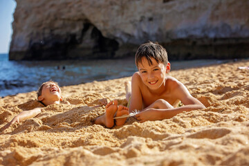 Child, tickling sibling on the beach on the feet with feather, kid cover in sand, smiling, laughing