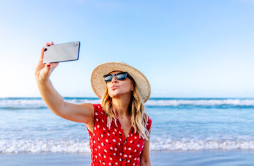 Blond woman wearing red dress and hat and using smartphone and taking a selfie at the beach, making...