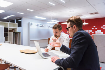 Two businessmen sitting in modern office, discussing in front of laptop