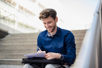 Smiling businessman sitting on stairs in the city taking notes