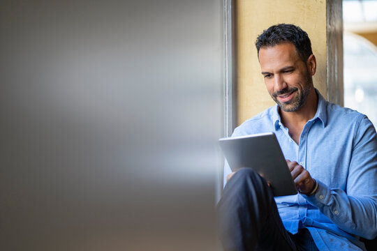 Smiling businessman sitting at the window using tablet