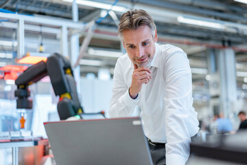 Confident businessman in a modern factory hall using laptop