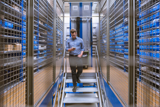 Businessman using smartphone walking down stairs in a factory