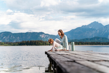 Mother with daughter sitting on jetty over lake against sky