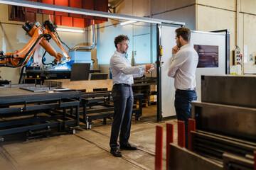 Young and mature businessman having discussion while standing against robotic machine at factory
