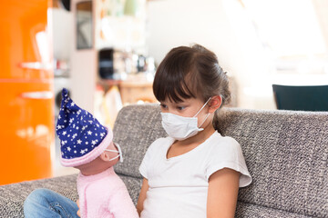Little girl with protective mask sitting on couch at home playing with doll
