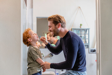 Happy man brushing son's teeth with toothbrush in bathroom at home