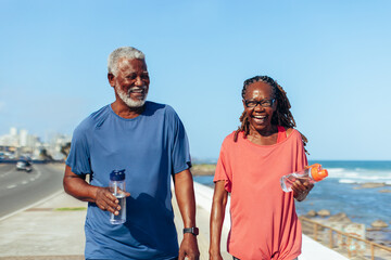 Joyful older couple taking a walk on the seaside promenade holding water bottles