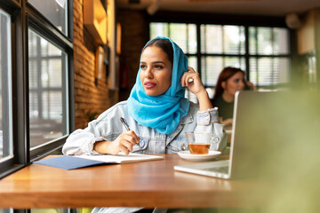 Businesswoman wearing turquoise hijab in a cafe and writing in notebook