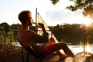 Young man with drinking a beer on a jetty next to sailing boat