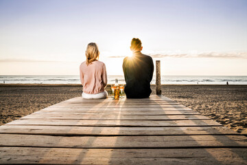 Couple with wine glasses sitting on boardwalk on the beach at sunset