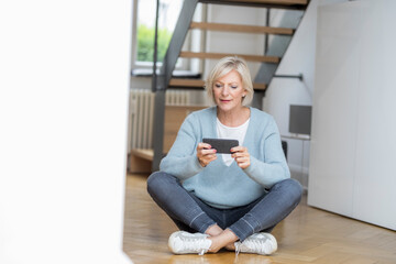 Portrait of senior woman sitting on the floor at home using smartphone