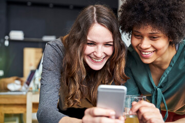 Two smiling young women sharing cell phone in office