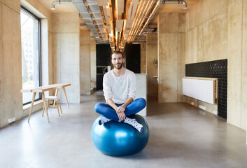 Portrait of young man sitting on fitness ball in modern office
