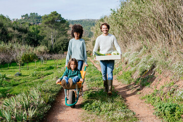 Family walking on a dirt track, pushing wheelbarrow, carrying crate with vegetables