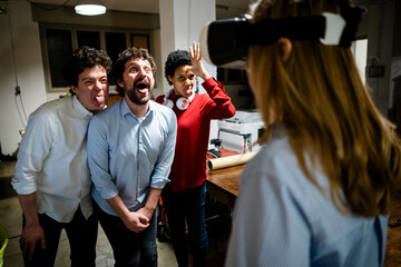 Playful colleagues looking at woman wearing VR glasses in office