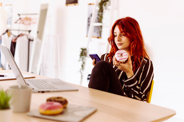 Young businesswoman holding doughnut while using mobile phone sitting at office