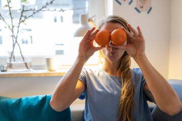Playful young woman covering her eyes with oranges at home