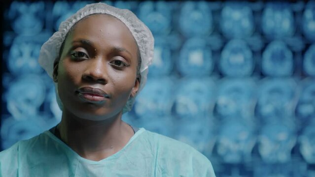 Chest up portrait of exhausted young Black female nurse taking off face mask and looking at camera standing in operating room against patient brain x-ray image on display