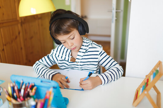 Boy doing homeschooling and writing on notebook, using tablet and headphones at home