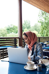 Young woman wearing hijab studying with her laptop on a terrace of a cafe