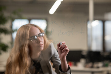 Caucasian woman with glasses writes text in English on a glass wall. 