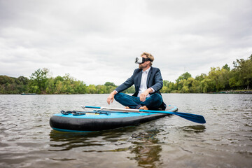Businessman wearing VR glasses sitting on SUP board on a lake