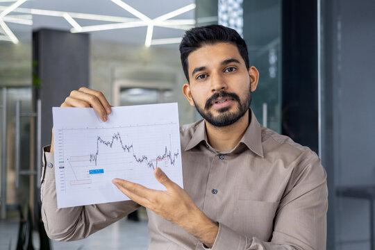 Professional businessman holding a financial chart, explaining market trends during a video call in a modern office setting.