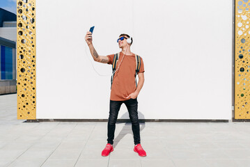 Smiling man wearing headphones taking selfie through mobile phone while standing with hands in...