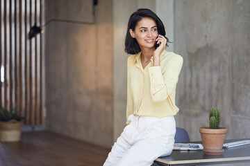 Smiling female entrepreneur talking over mobile phone while sitting on desk in office