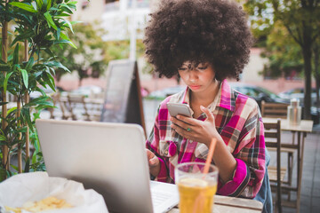 Young woman with afro hairdo using smartphone and laptop at an outdoor cafe in the city