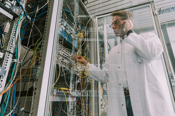 Mature man talking over smart phone while plugging transceiver on fiber optic cable in data center